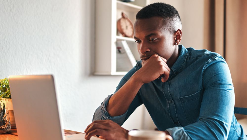 Man in denim shirt, focused, using a laptop, resting chin on hand, looking at screen. Indoor setting.