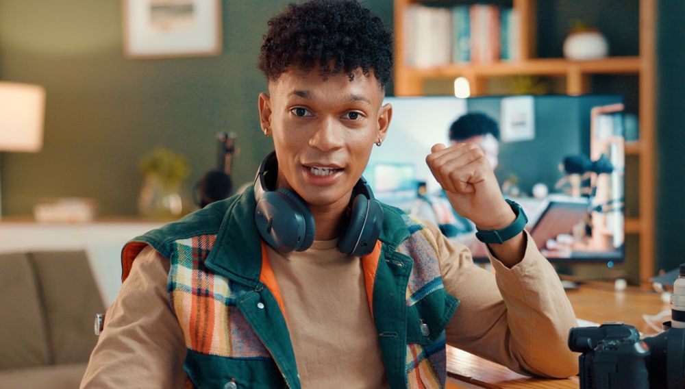 Young person with headphones, fist raised, sitting at a desk with camera and computer.