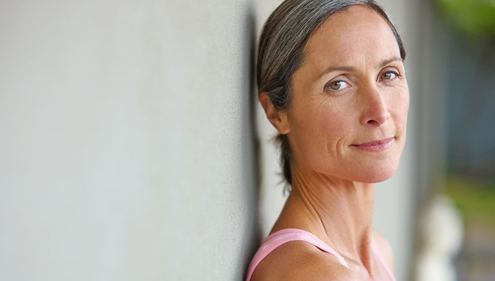 Woman with graying hair leans against a wall, smiling softly.
