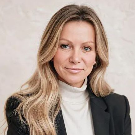 Woman with blonde hair wearing a black blazer and white turtleneck, looking at the camera.