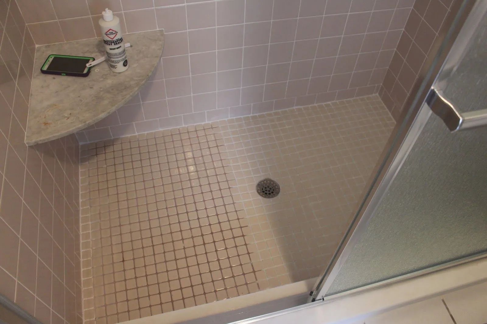 Pink-tiled shower stall with a gray corner shelf holding a phone and cleaning supplies. A shower door is on the right.