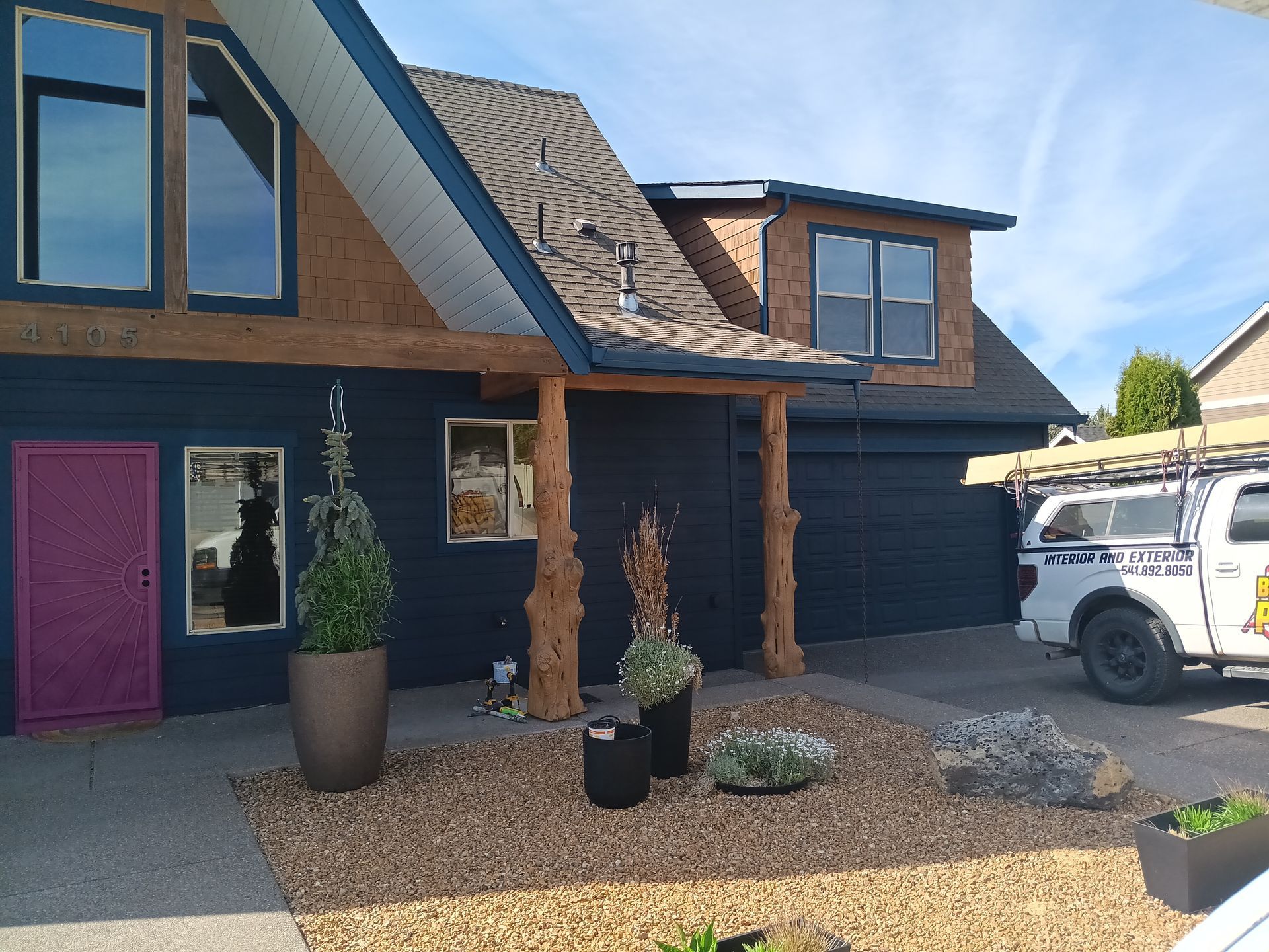 Modern dark-blue house exterior with pink door, wood posts, gravel yard, and white work van parked outside