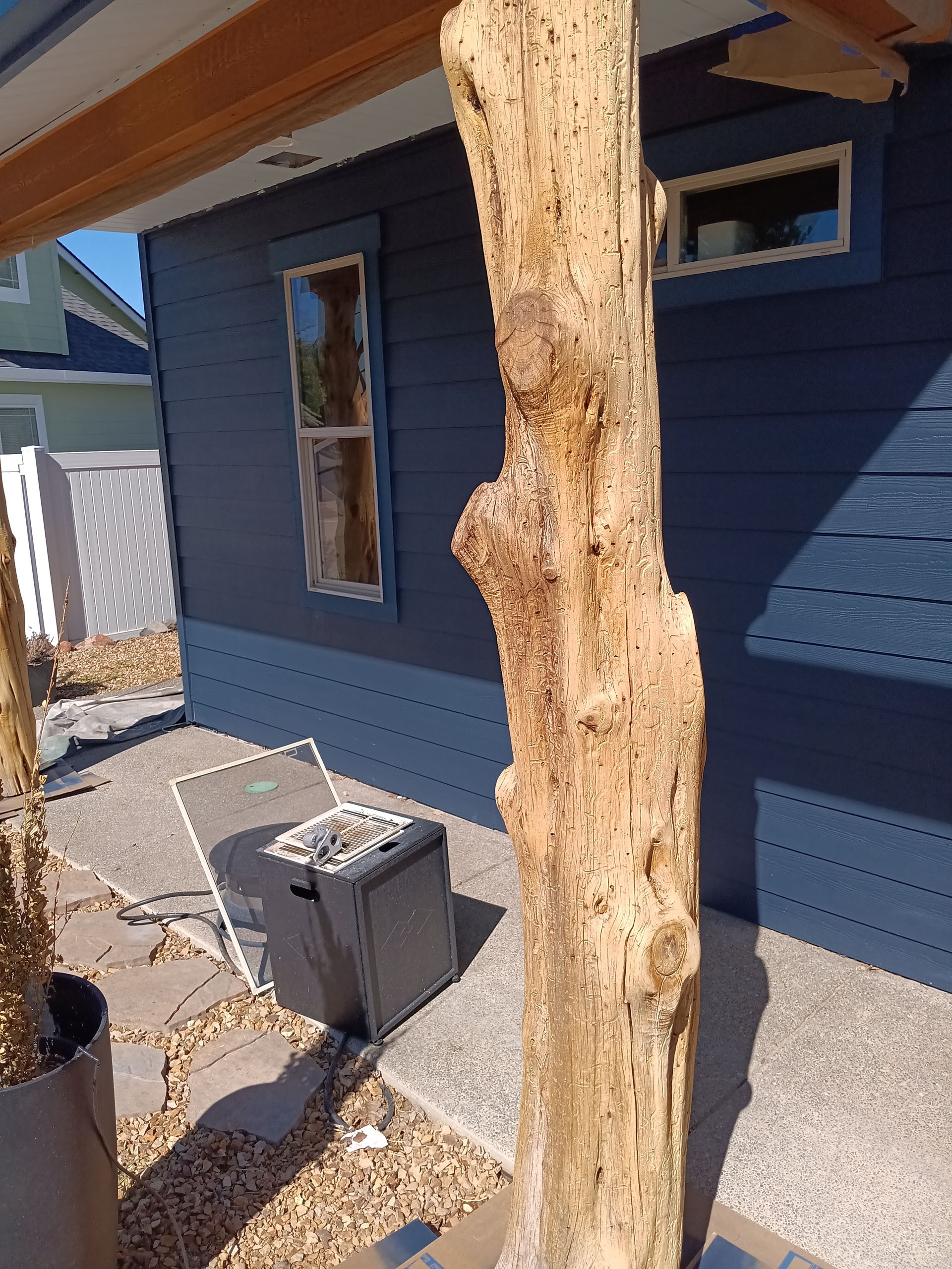Juniper tree trunk sealed; supports the balcony besides a beside a blue house with a small window and a trash bin on a concrete patio.