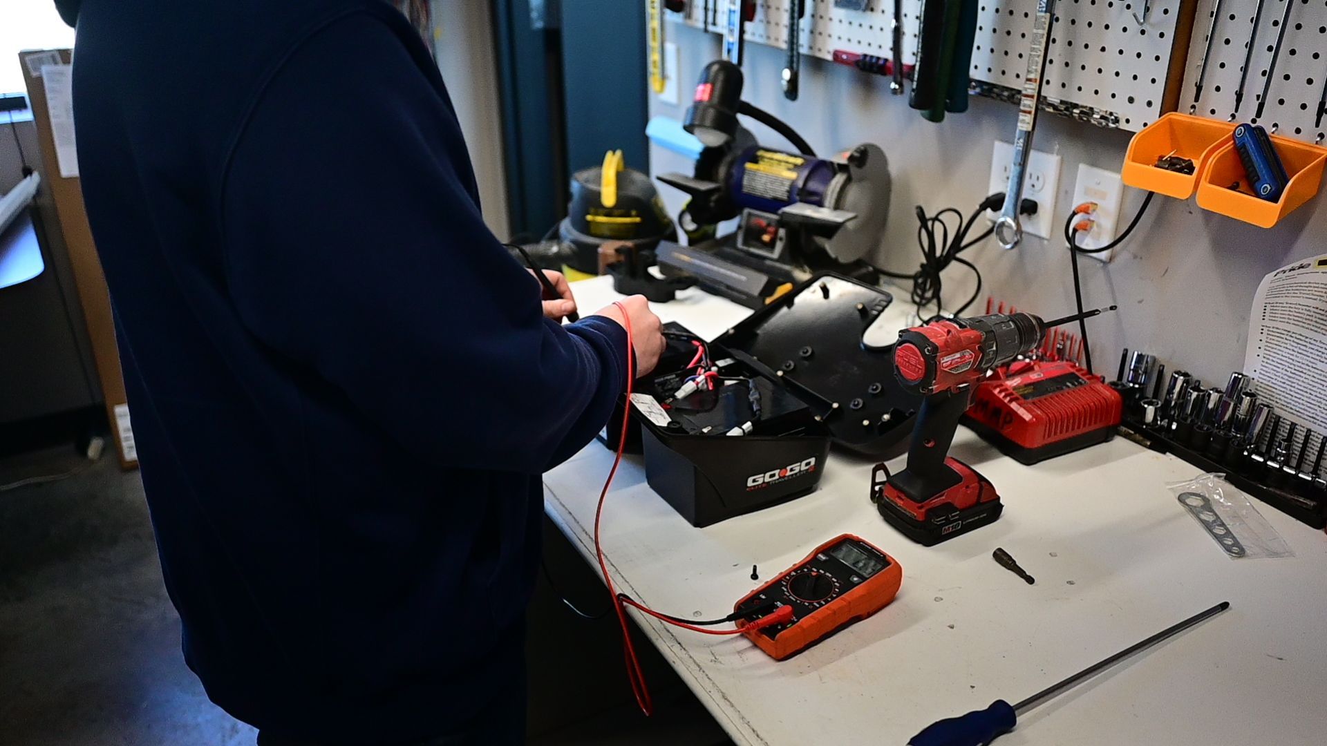 Person using a multimeter on a device in a workshop. Tools and equipment are on the workbench.