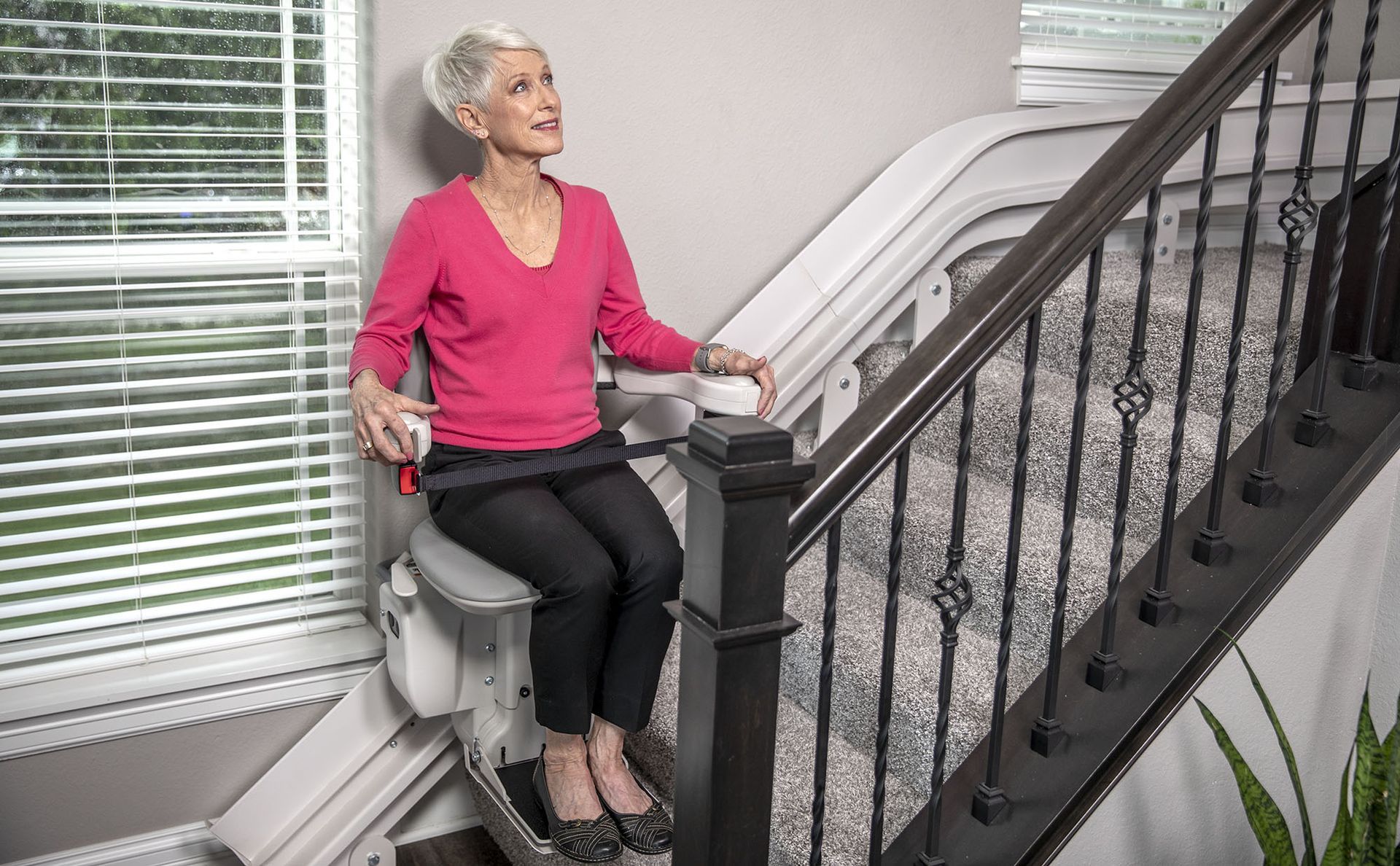 Woman on stairlift, pink top, black pants, going up stairs, window to the left.