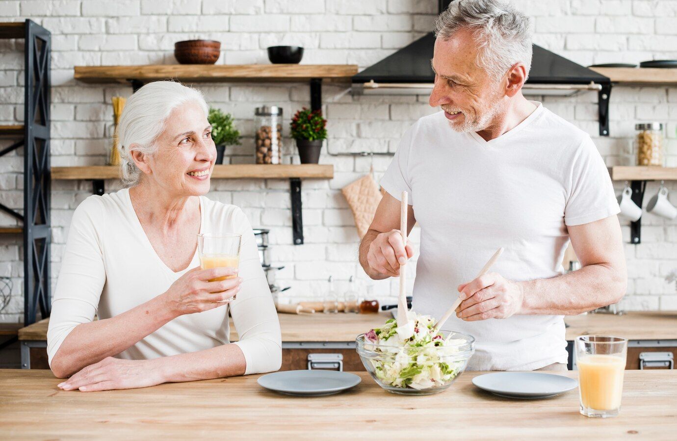 An elderly couple is sitting at a table in a kitchen eating a salad.