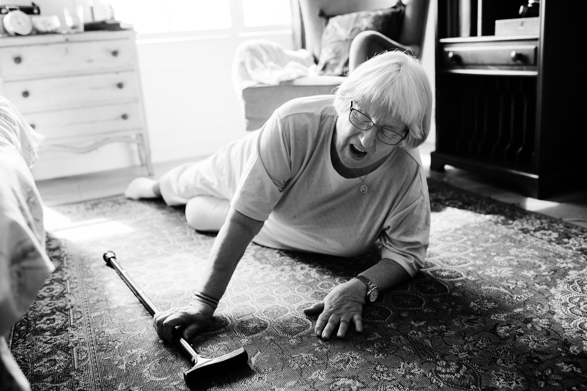 Elderly woman lying on the floor after a fall, cane nearby, in a bedroom.