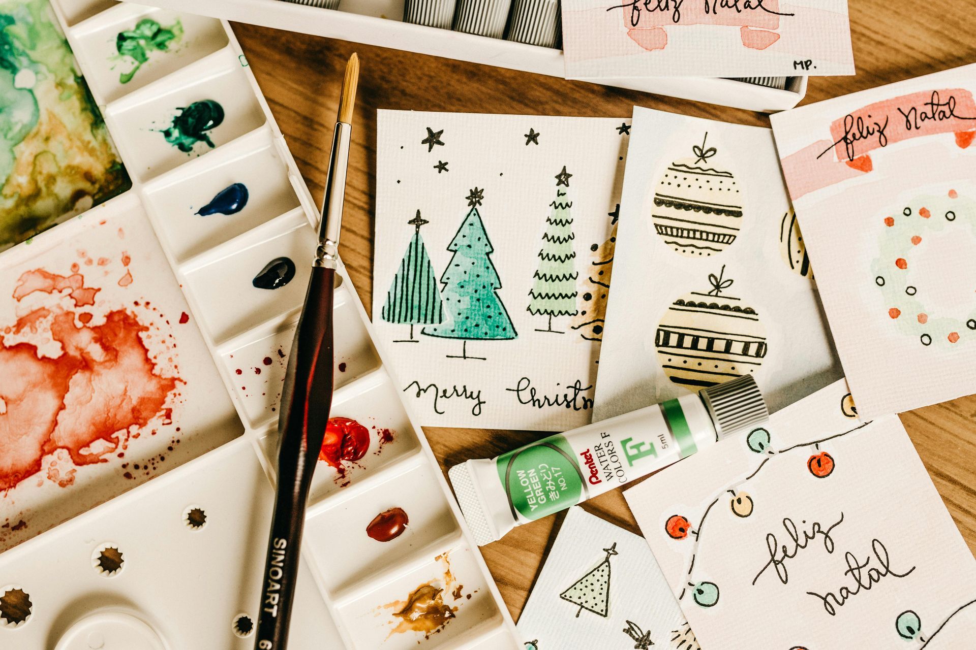 Watercolor Christmas cards and supplies on a wooden surface: paint palette, brush, green paint tube, and cards.
