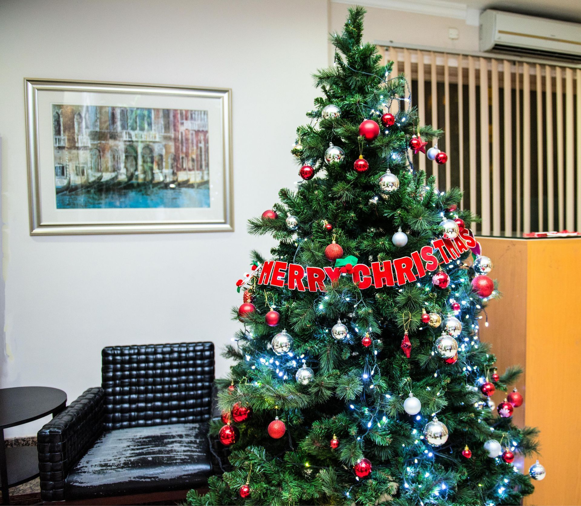 Christmas tree decorated with ornaments and lights, next to an armchair and painting.