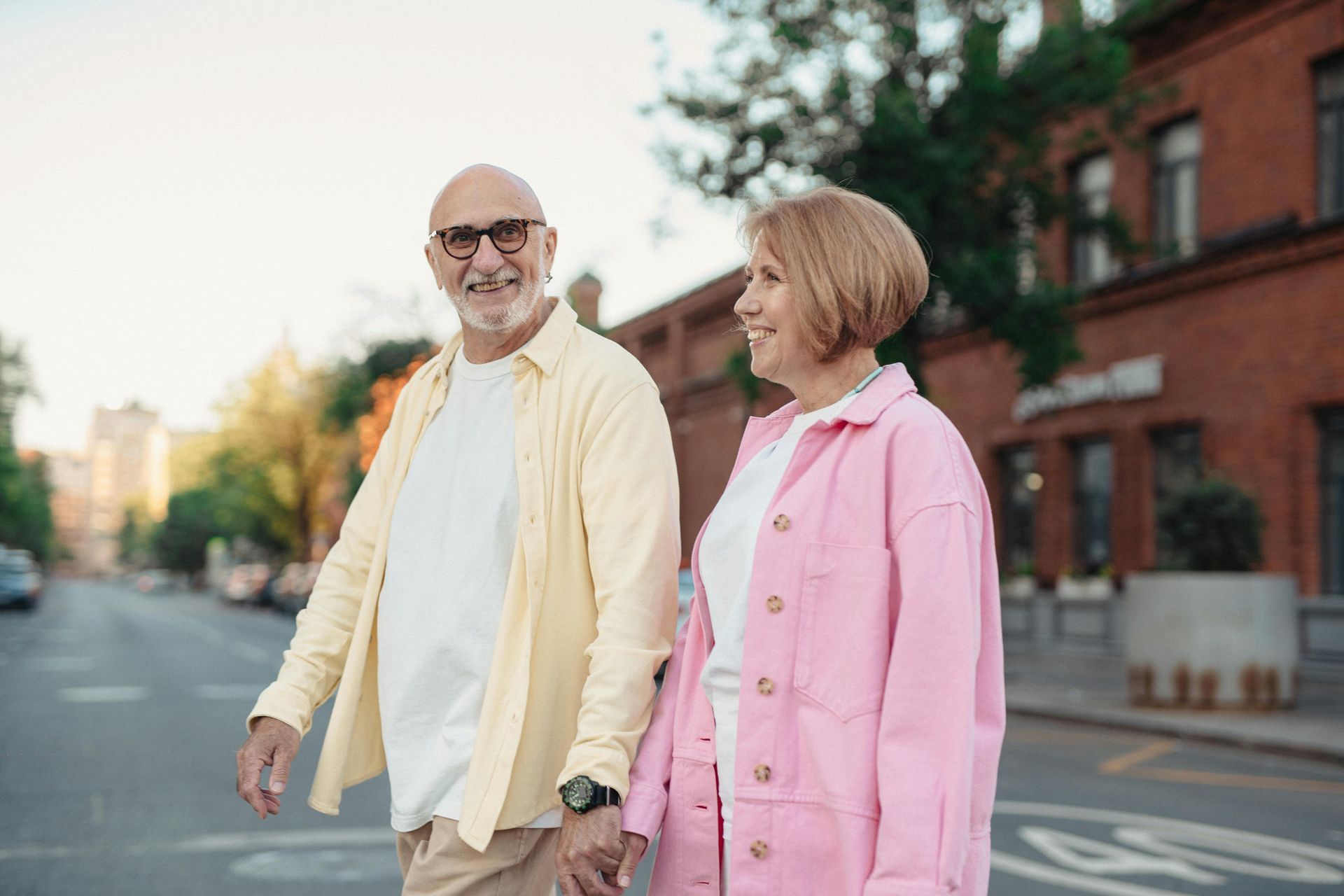 Elderly couple holding hands, crossing a street. Man in yellow shirt, woman in pink, both smiling. City background.