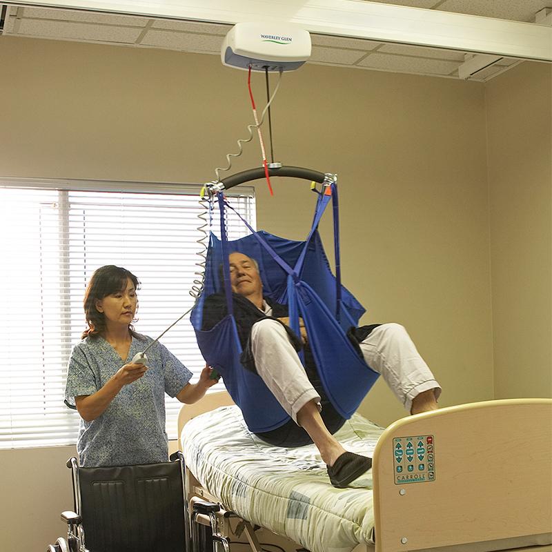 Woman assisting a person with a ceiling lift, blue sling, in a hospital room.