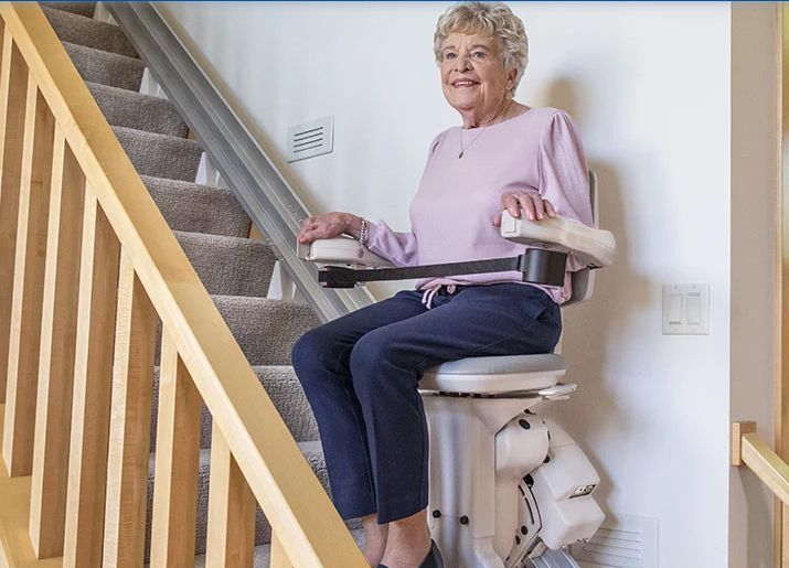 Woman using a stairlift to ascend a carpeted staircase. She smiles, holding the armrests.