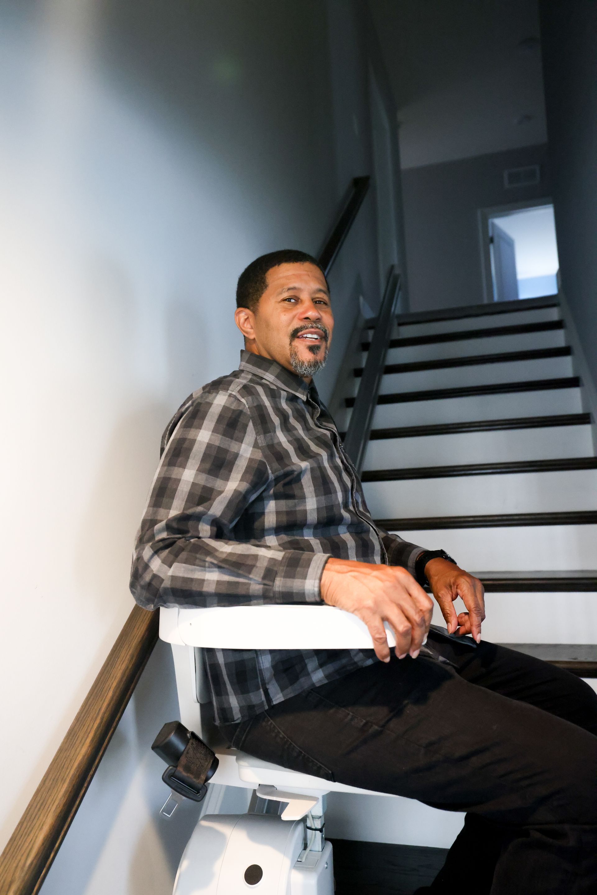 Man smiling on a stair lift going up a staircase. He wears a plaid shirt and black pants.