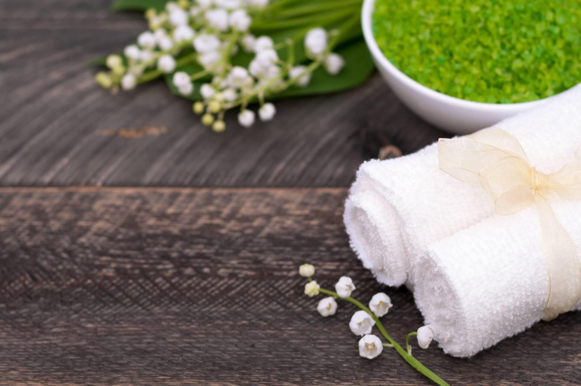 Rolled white towels tied with a ribbon, bowl of green bath salts, and lily of the valley flowers on a wood surface.