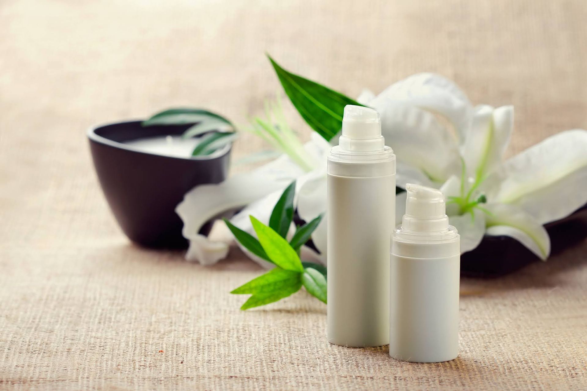 Two white skincare bottles with a lily and bowl of cream on a beige cloth.