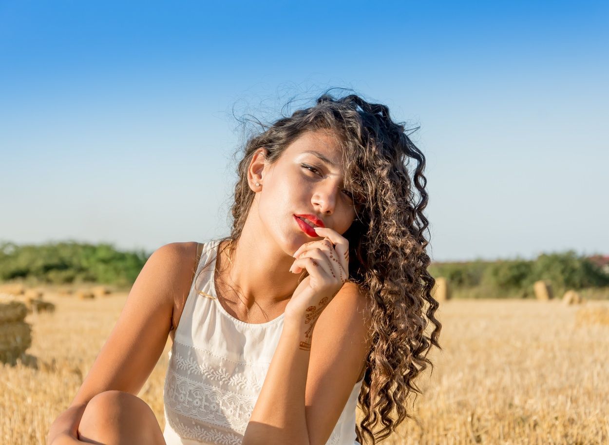Woman with curly hair in a white dress, holding a red berry, sitting in a wheat field under a blue sky.