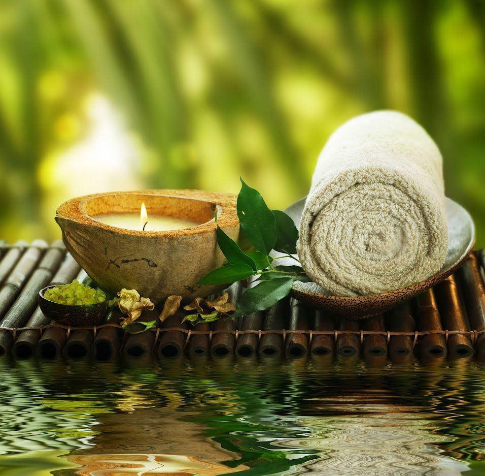 Spa still life: lit candle in coconut shell, rolled towel, green leaves, bamboo mat reflected in water.