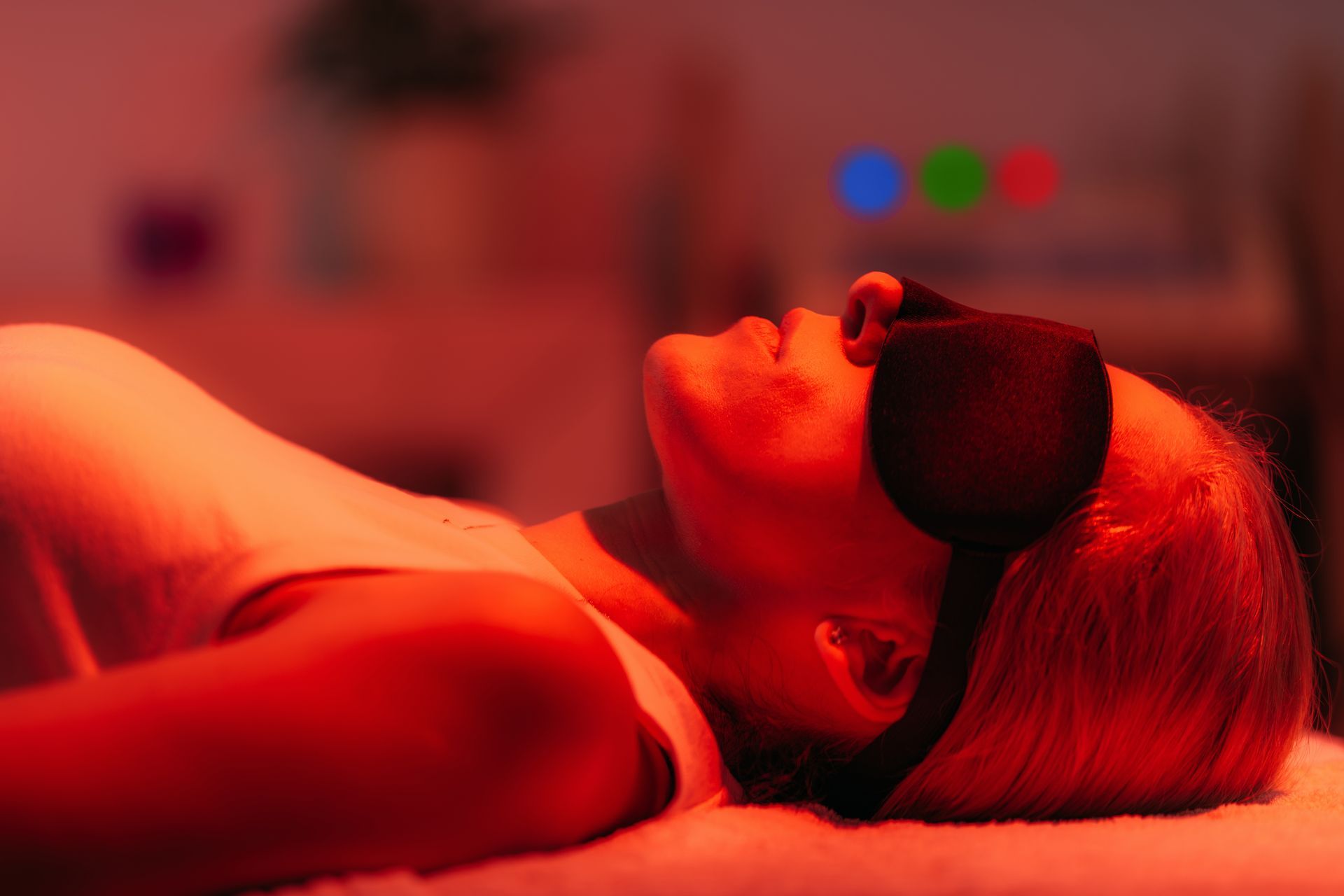 Person wearing an eye mask relaxes under red light therapy in a dim treatment room. Person wearing an eye mask relaxes under red light therapy in a dim treatment room.