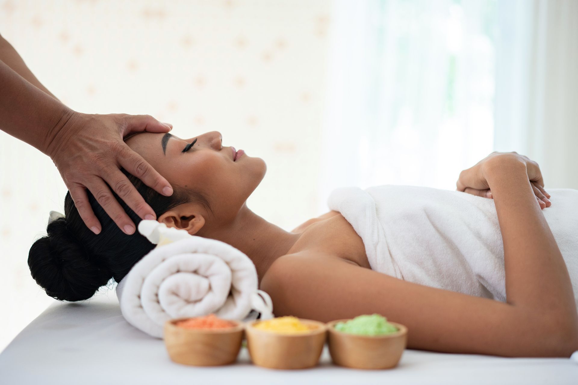 Woman receiving head massage at a spa; eyes closed, towel, and colorful bowls.