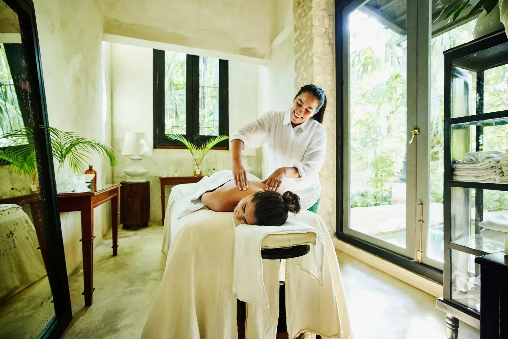 Woman receiving massage in a bright spa room; therapist applying pressure to back.