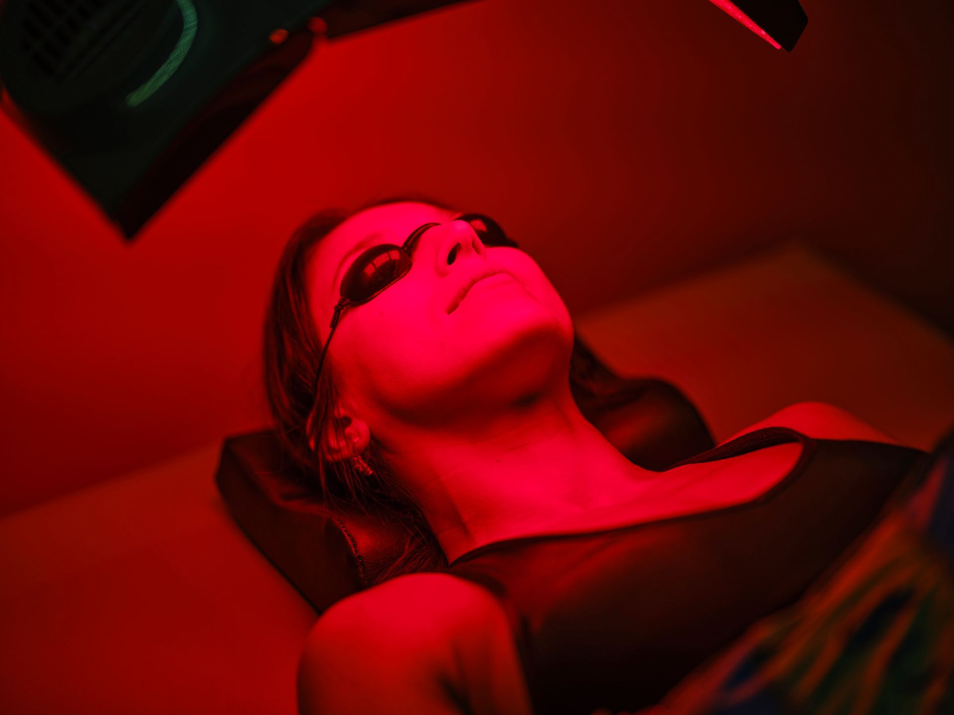 Woman wearing eye protection lies under red light therapy, lit in red.