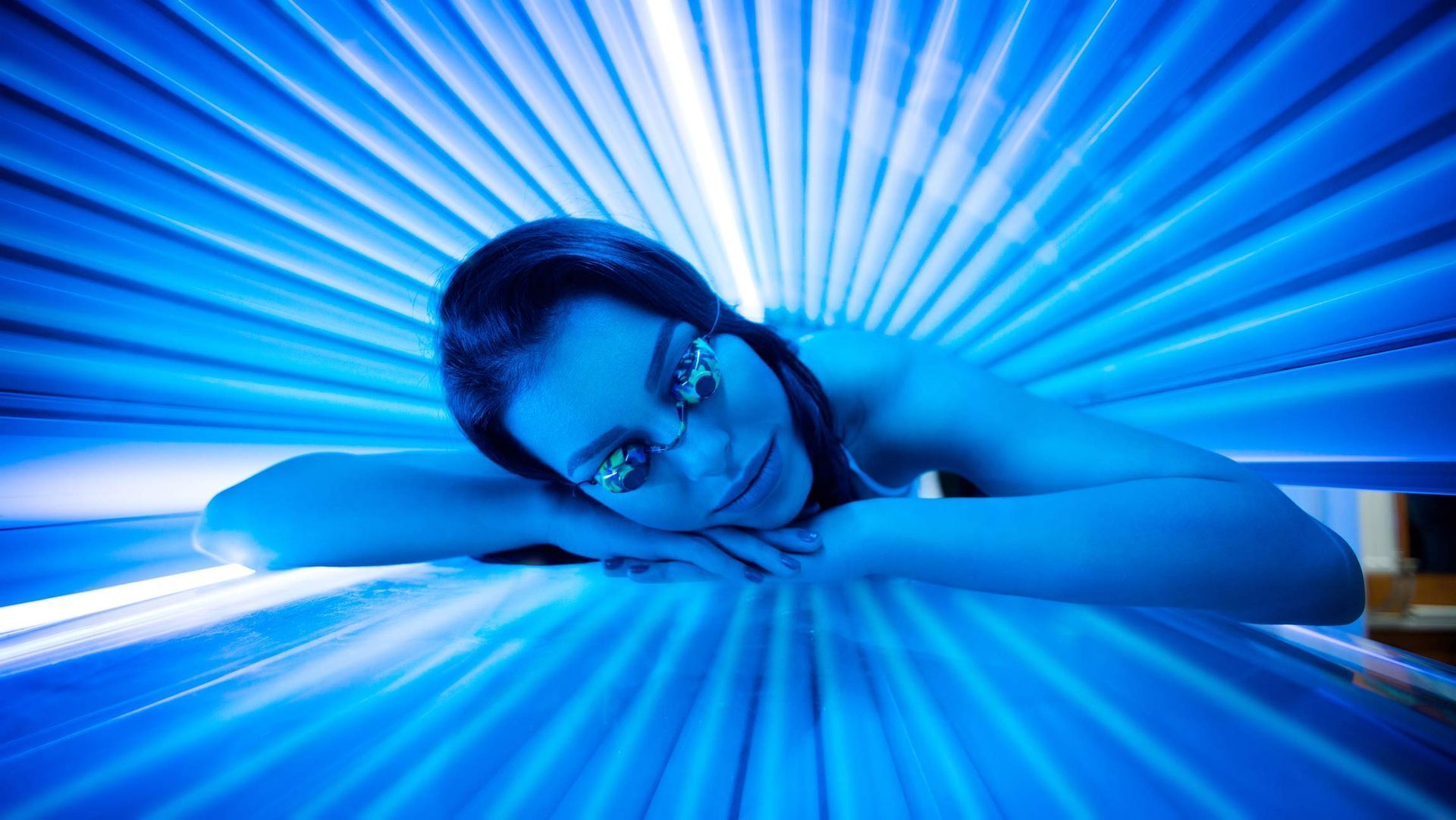 Woman in tanning bed, eyes covered, bathed in blue light.