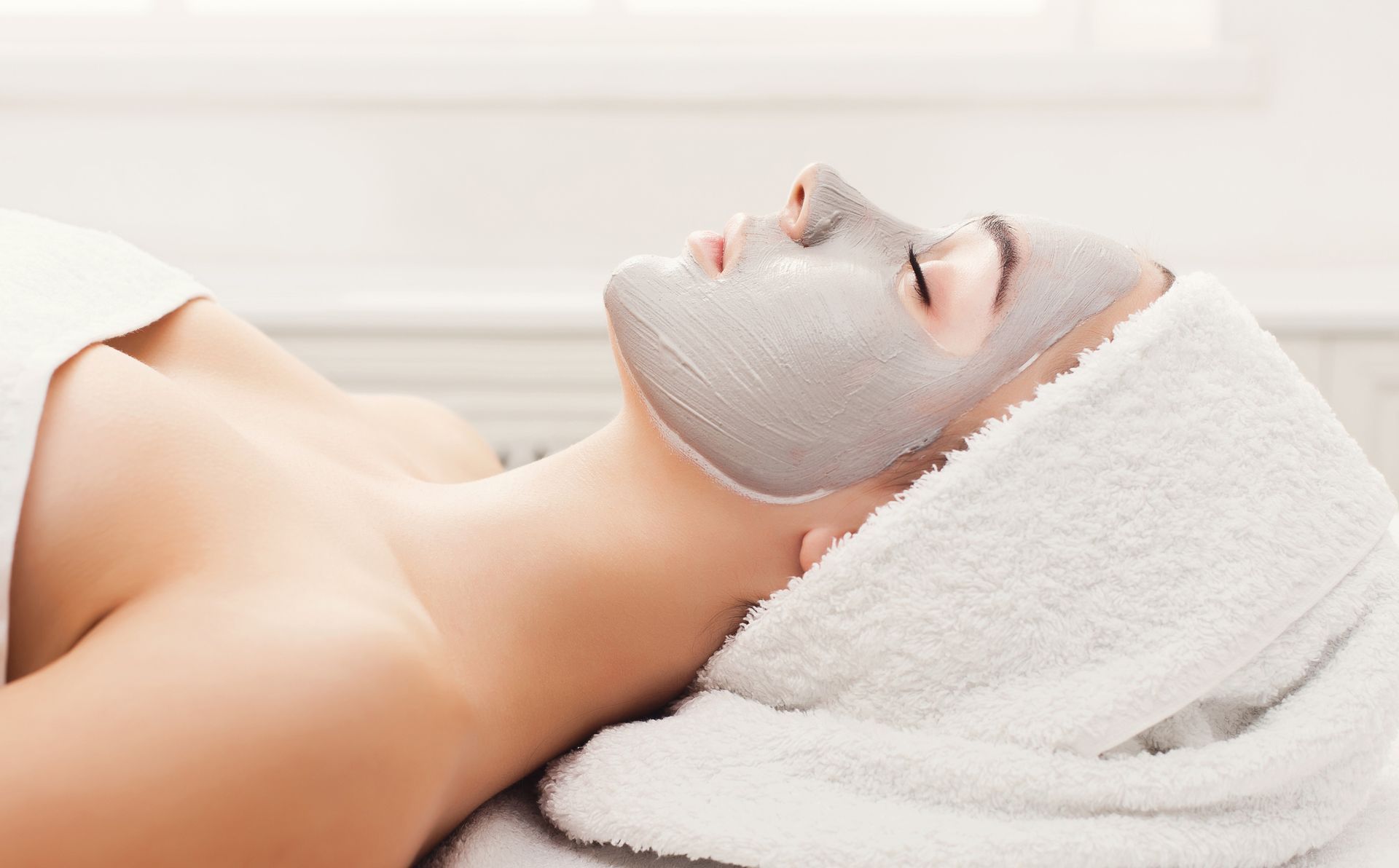 Person with facial mask, relaxing with eyes closed, wrapped in a white towel, spa setting.