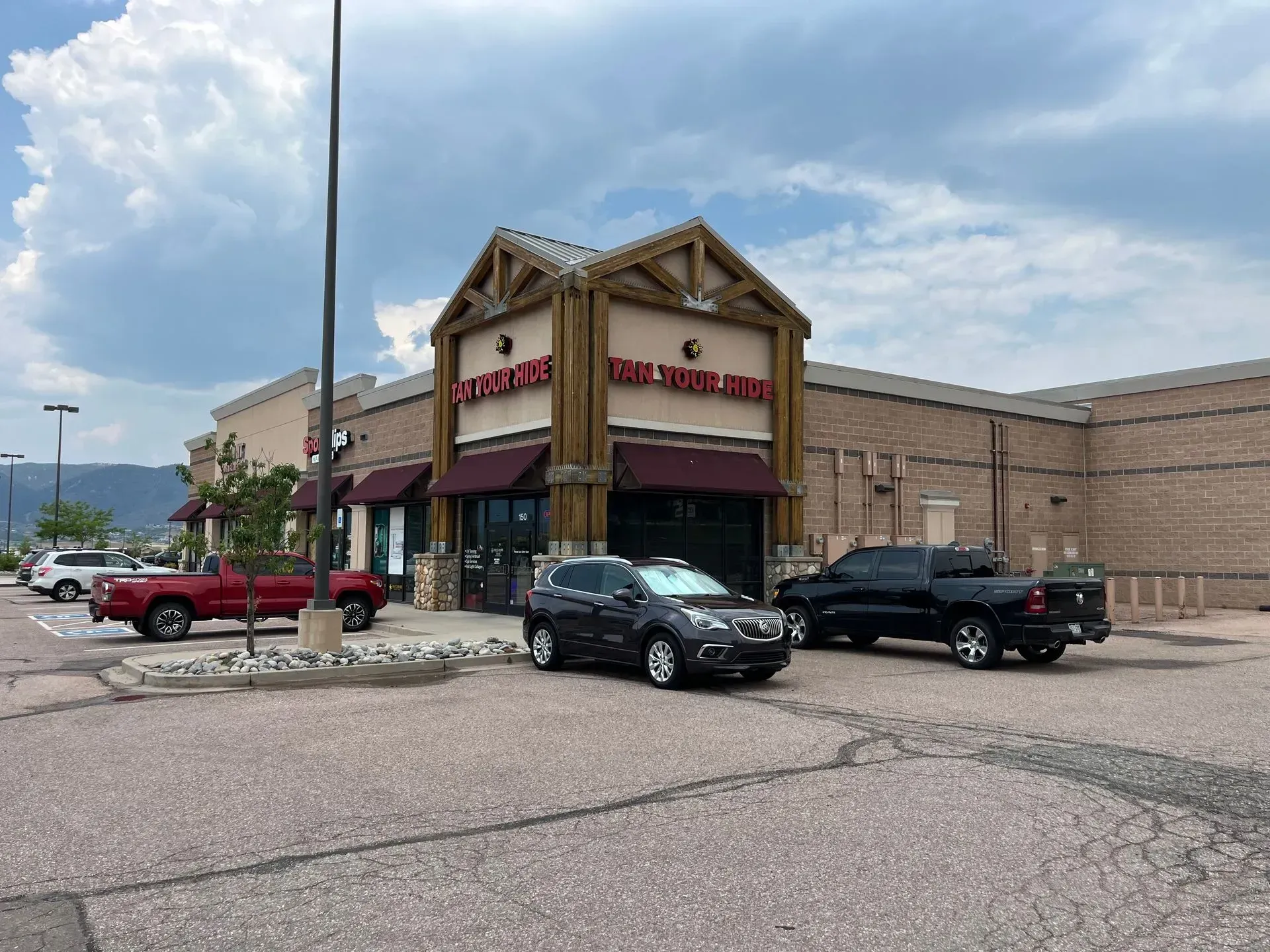 Exterior view of a shopping center with a restaurant, cars parked in the lot, and mountains in the background under a cloudy sky.
