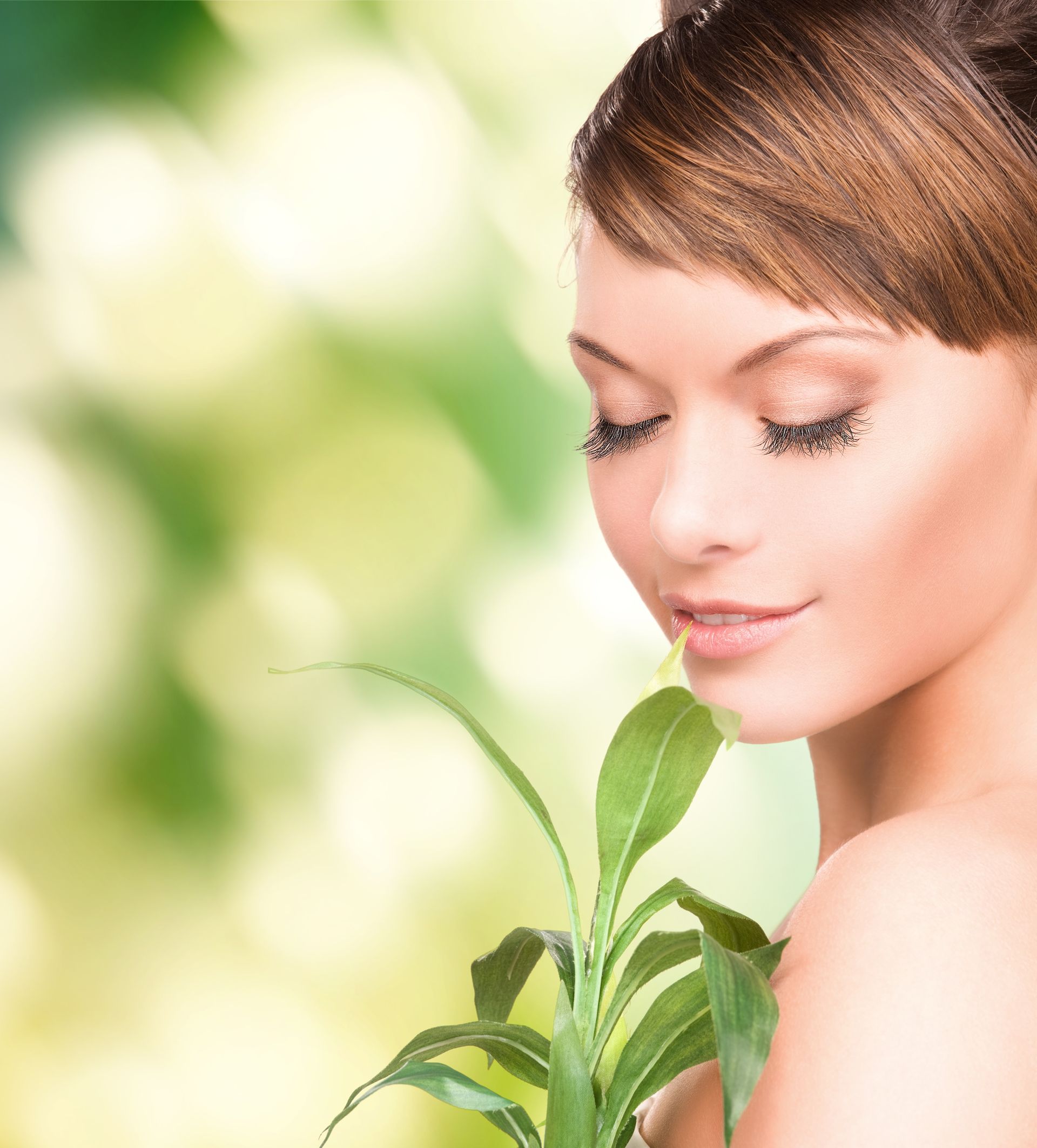 Woman with brown hair holding a green plant, eyes closed, against a blurred green background.
