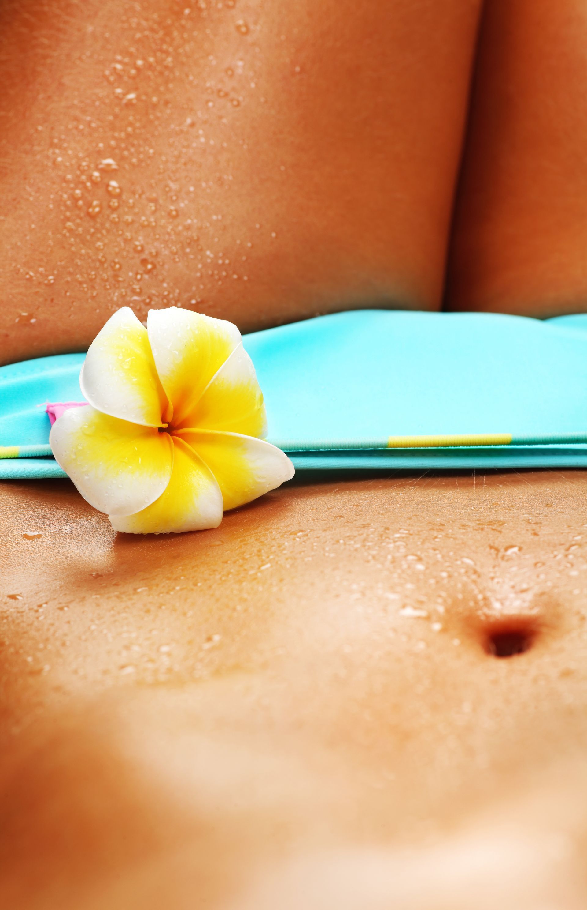 Close-up of a person's torso. Turquoise bikini top, a flower rests on the skin, and water droplets.
