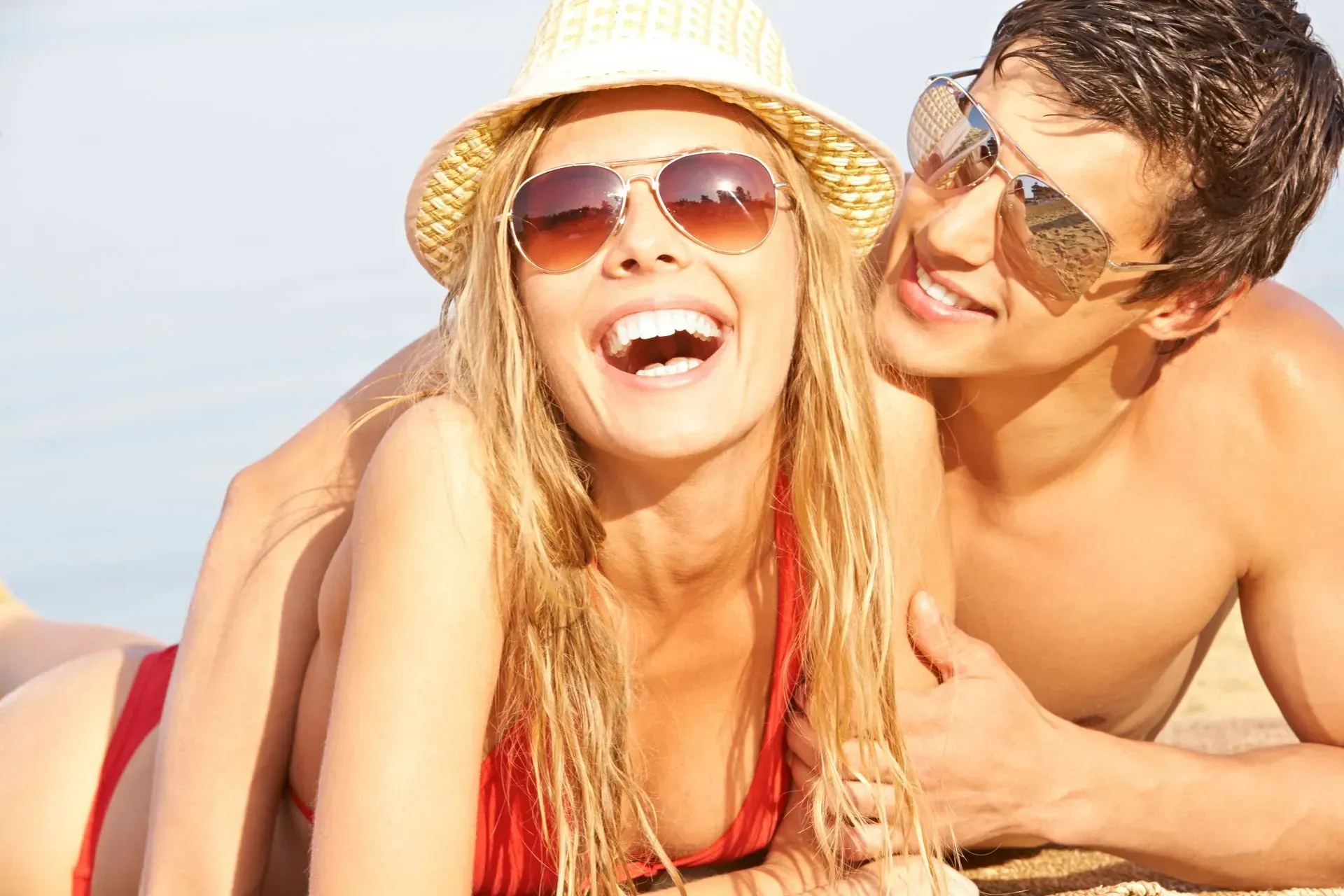 Woman in red bikini laughing; man smiles, sunglasses; outdoors, sunny.