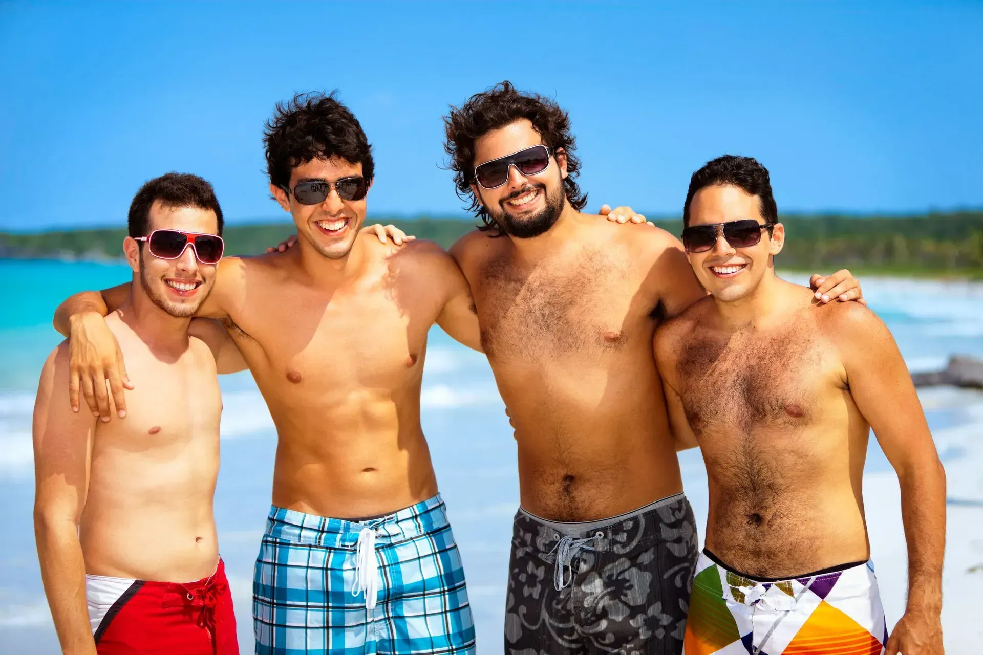 Four shirtless men with sunglasses, arm-in-arm, smiling on a beach.