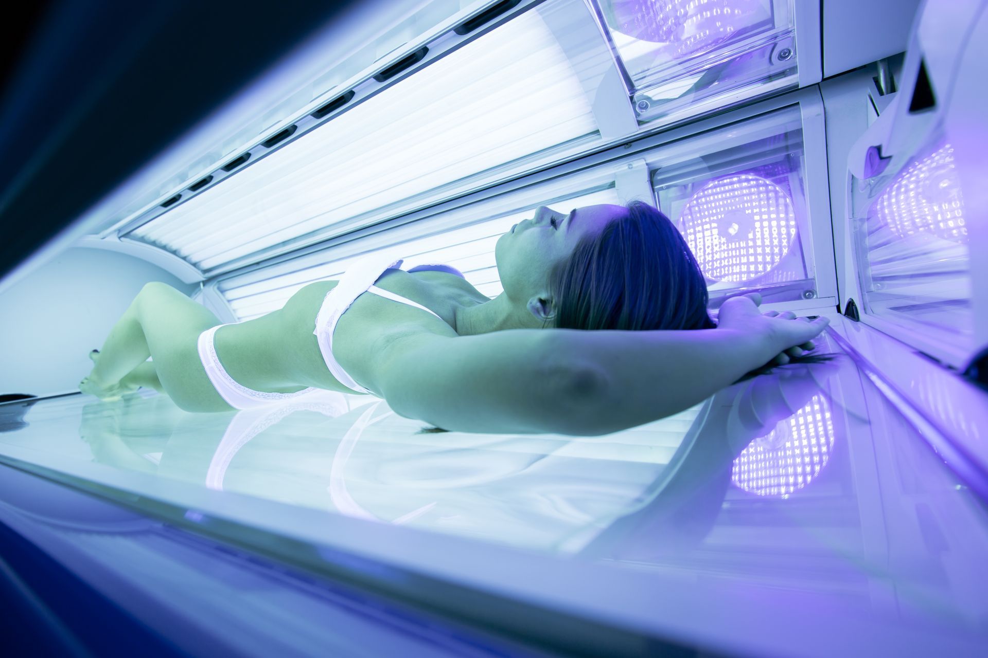 Woman in a tanning bed, lying down with eyes closed, exposed skin, surrounded by blue light.