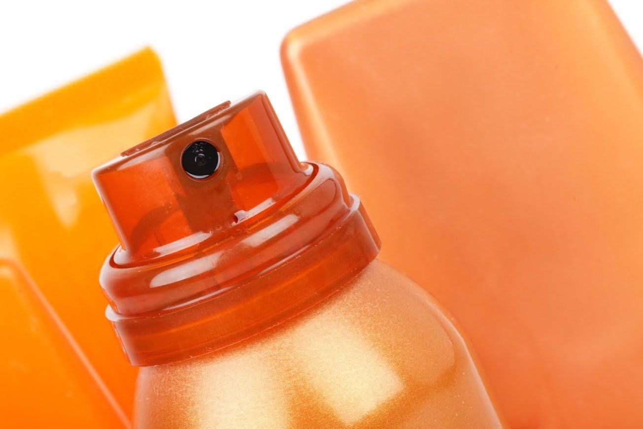 Close-up of a spray sunscreen bottle, orange-tinted with nozzle detail, blurred orange background.