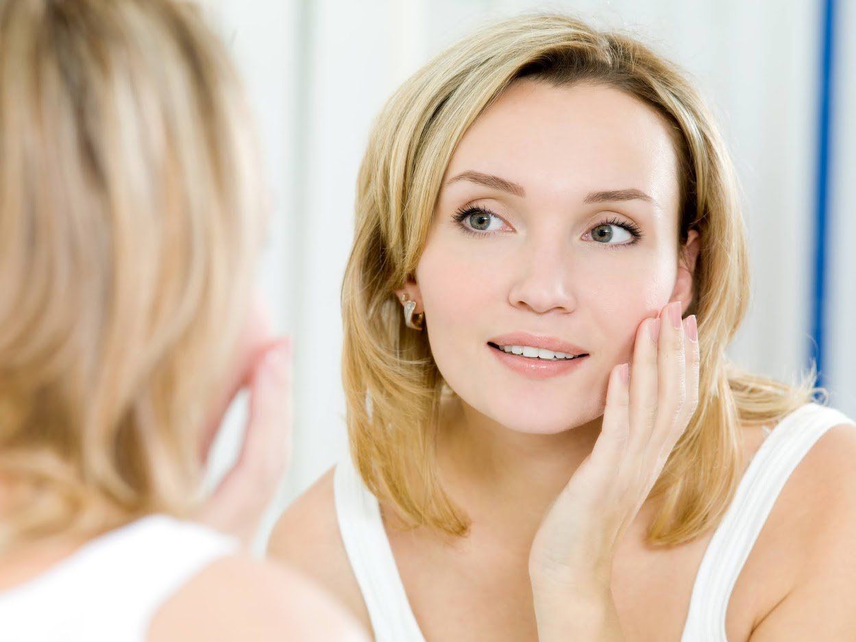 Woman looks in mirror, touching her cheek with a smile, light hair, white top, bright setting.