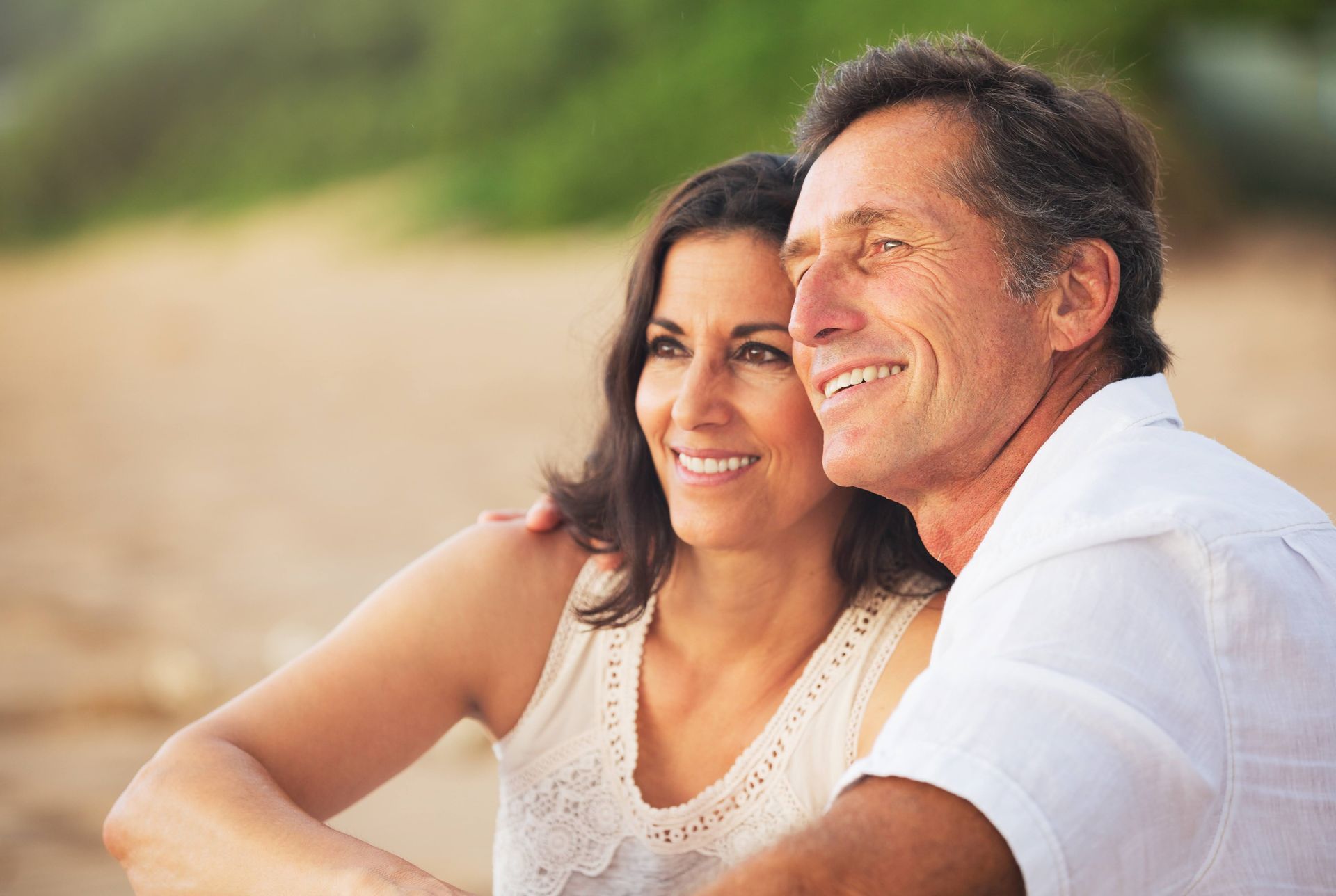 Couple smiling, looking off-camera on a beach. Woman's arm around man's shoulder. Natural light.