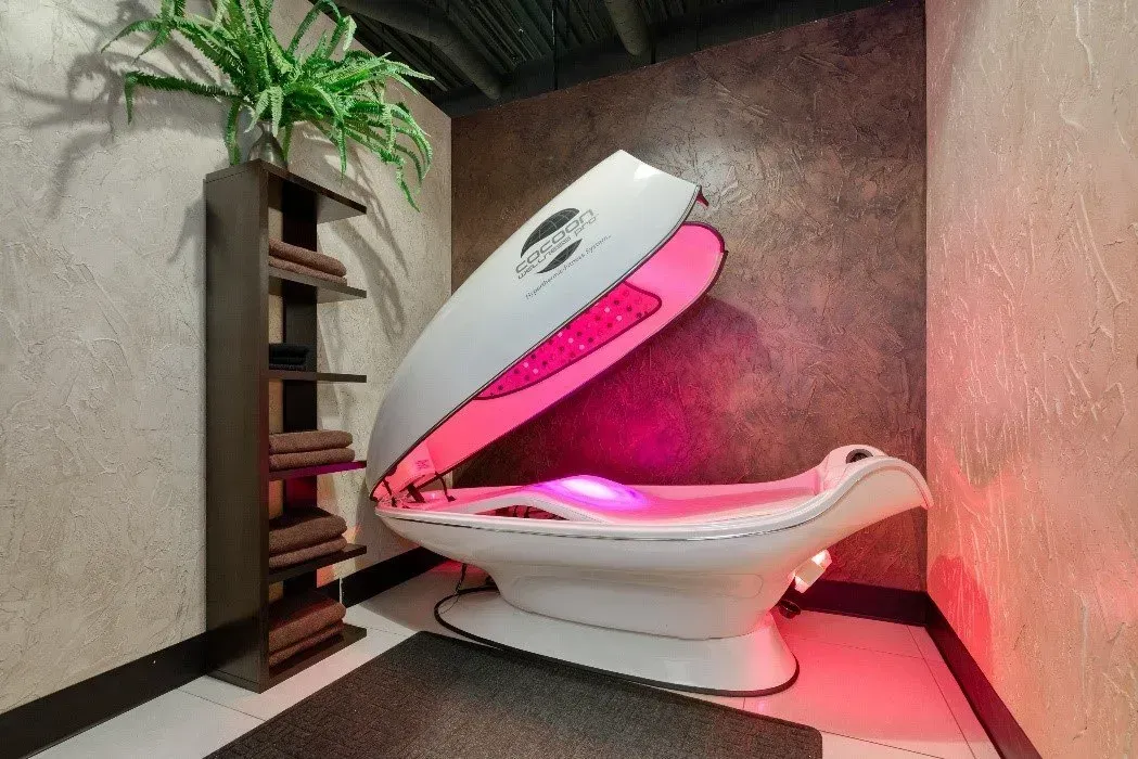 A white spa pod with pink interior, against a textured wall. A plant and shelves are nearby.