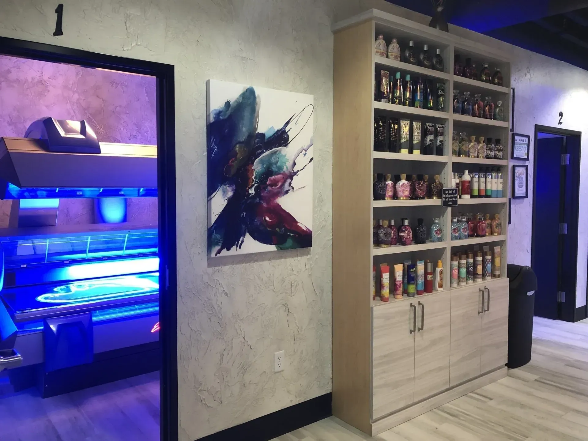 Tanning salon interior with open tanning bed, lotion display, and abstract artwork on the wall.