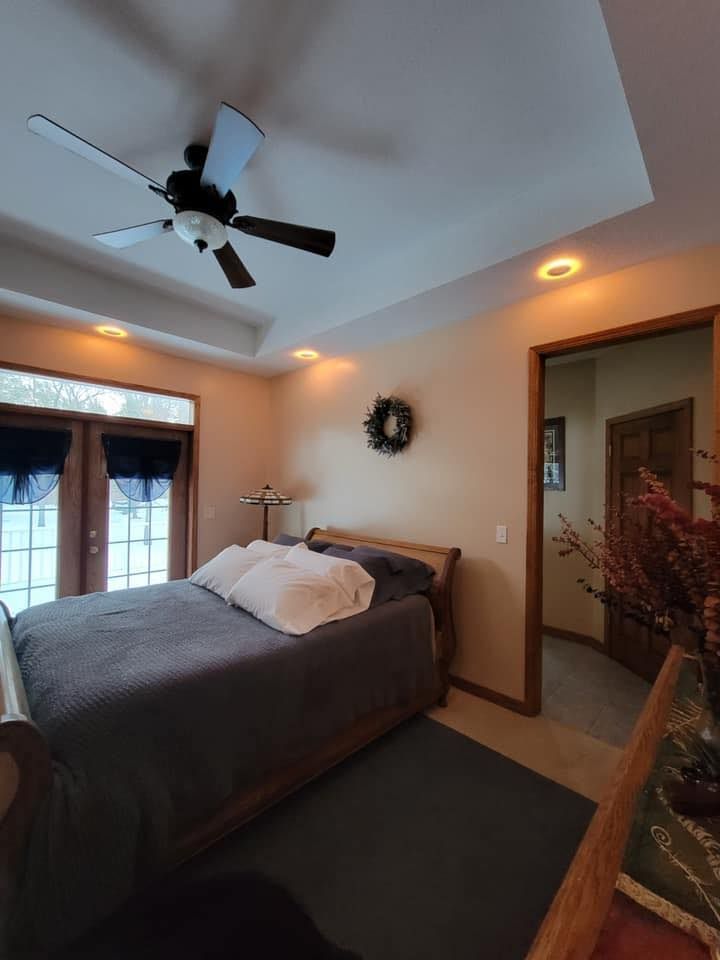 A cozy bedroom with a queen bed, gray bedding, wooden headboard, ceiling fan, and a doorway leading to a hallway.