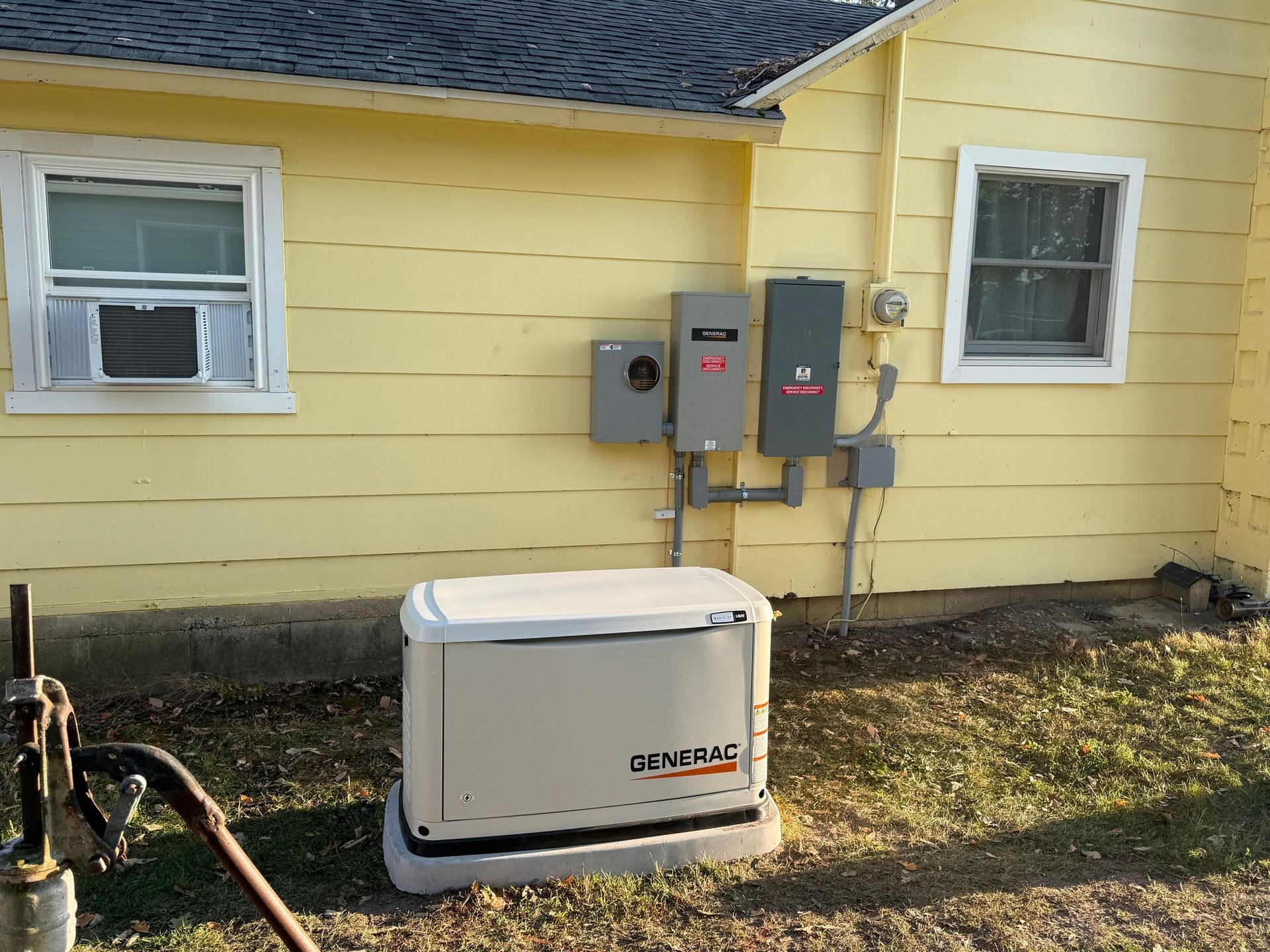 A Generac backup generator sits on the ground outside a yellow house with electrical boxes mounted on the wall.
