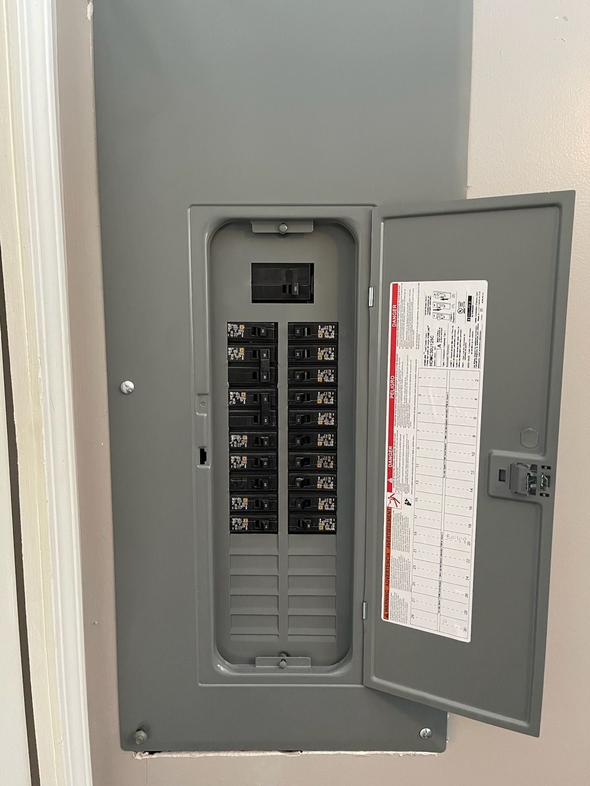 A gray indoor electrical circuit breaker panel mounted on a wall, with its door open to reveal multiple circuit switches.