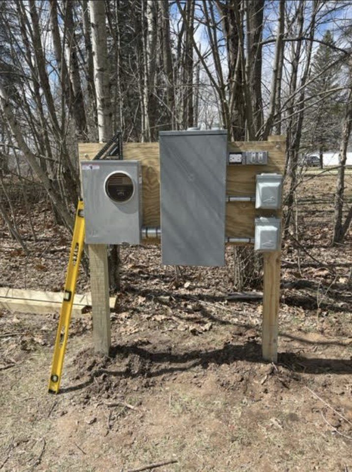 A gray electrical meter base and service panel mounted on a wooden frame outdoors with a yellow level leaning against it.