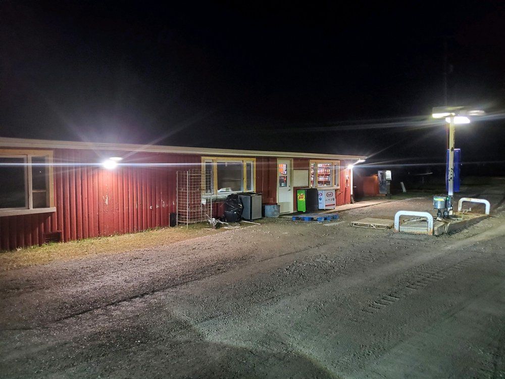 A small red building with bright outdoor lights at night, featuring a gravel area with metal parking barriers in front.