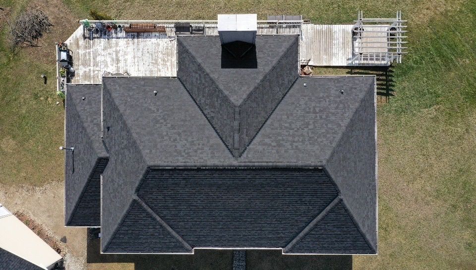 An aerial, top-down view of a dark-shingled residential roof with a central chimney, adjacent to a patio and grassy yard.