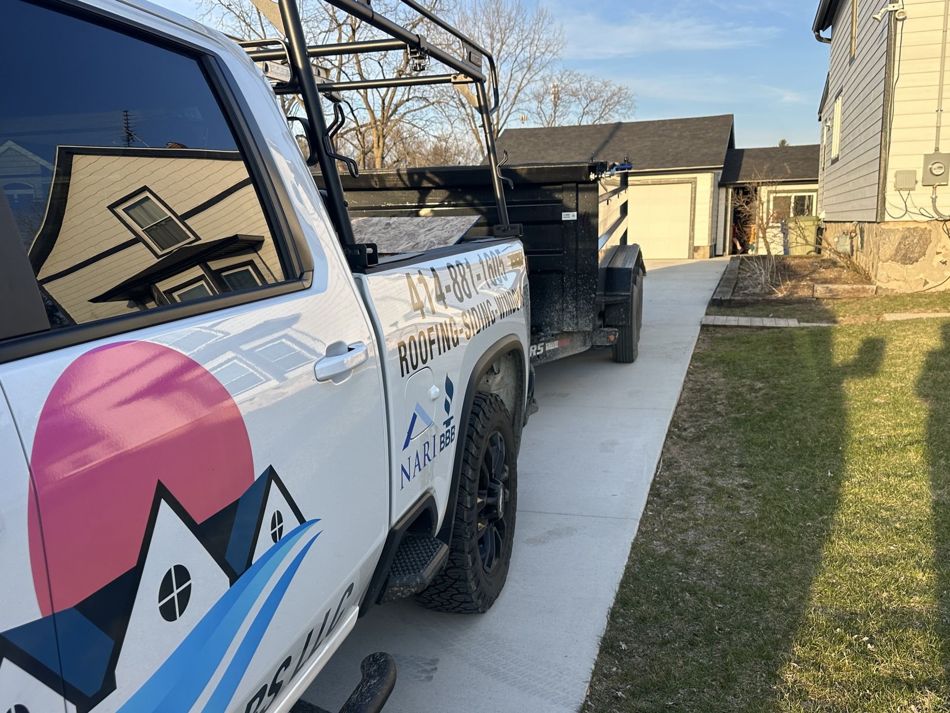 A white pickup truck with a logo and a trailer attached, parked on a residential concrete driveway.