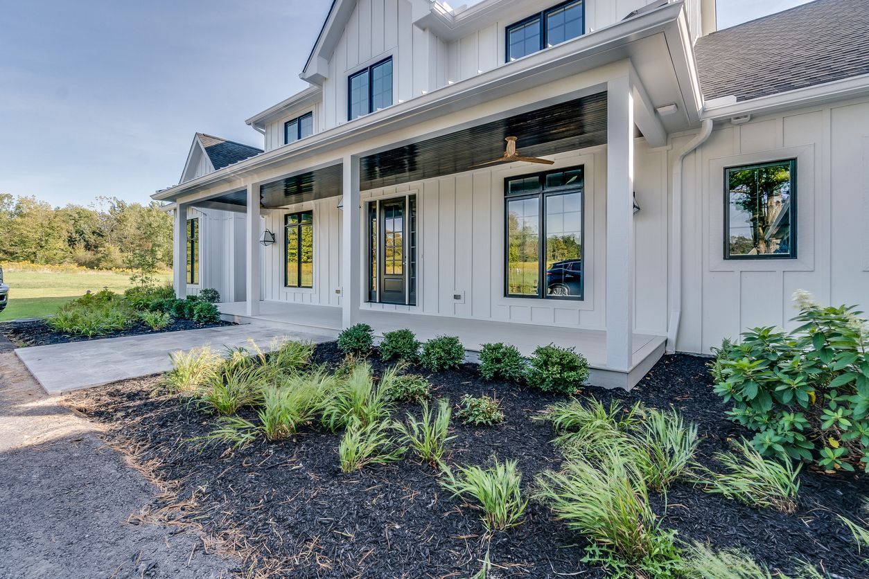 White modern farmhouse with black-trimmed windows, a covered front porch, and landscaped garden beds on a sunny day.