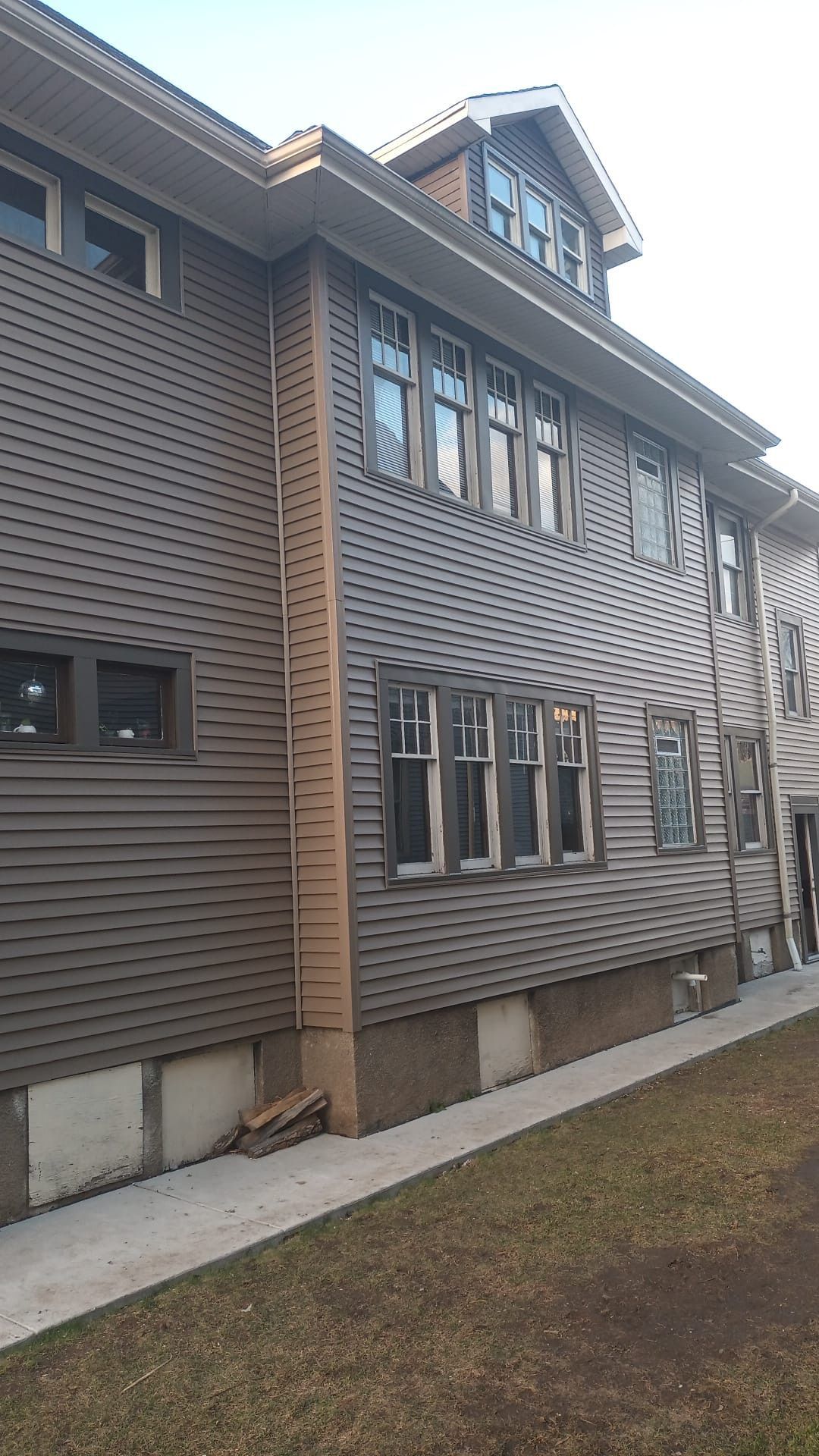 Side view of a brown two-story house with horizontal siding, dark-framed windows, and a concrete foundation.