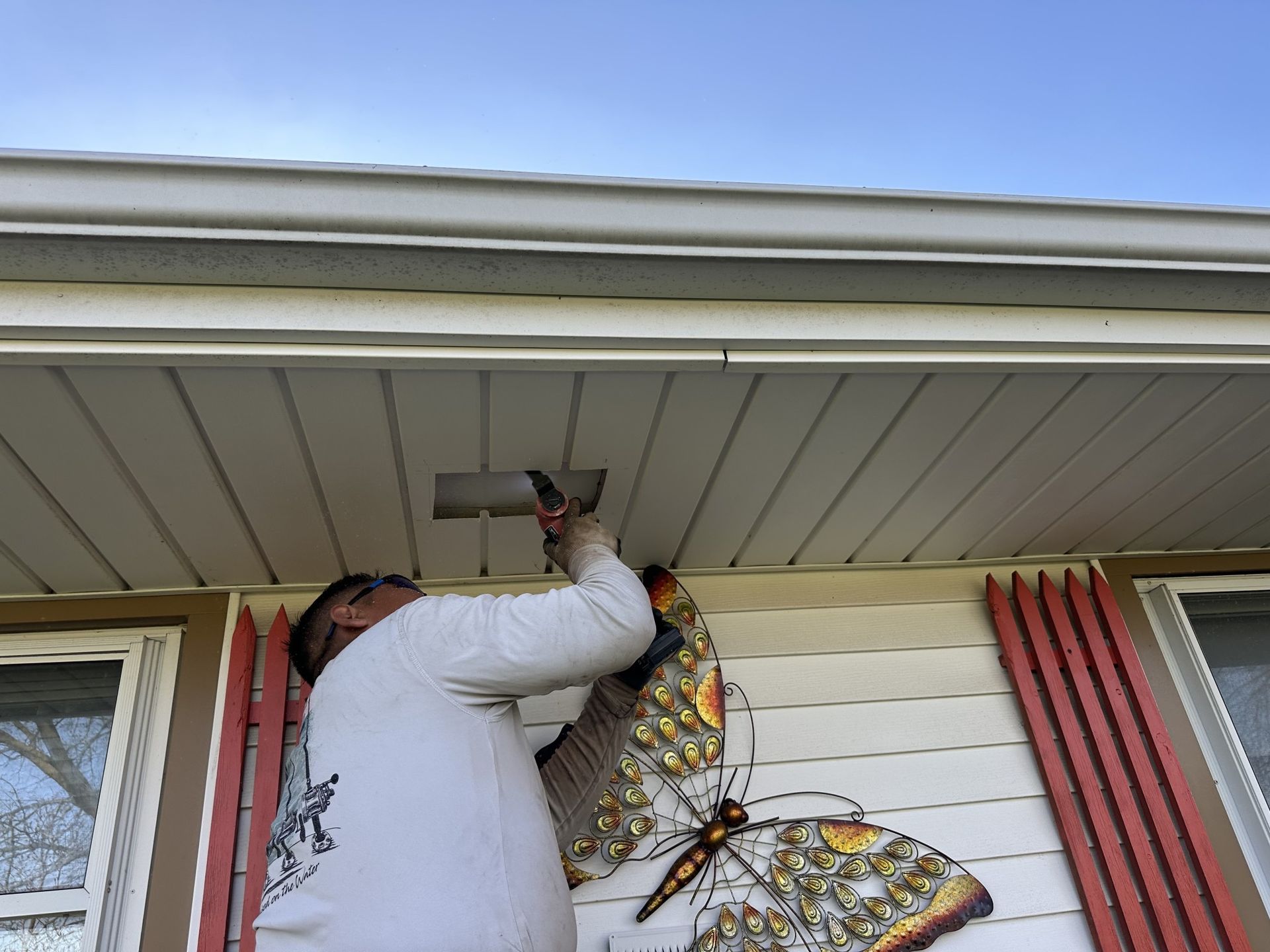 A person stands on a ladder working on an exterior soffit light fixture on a house with white siding and red shutters.