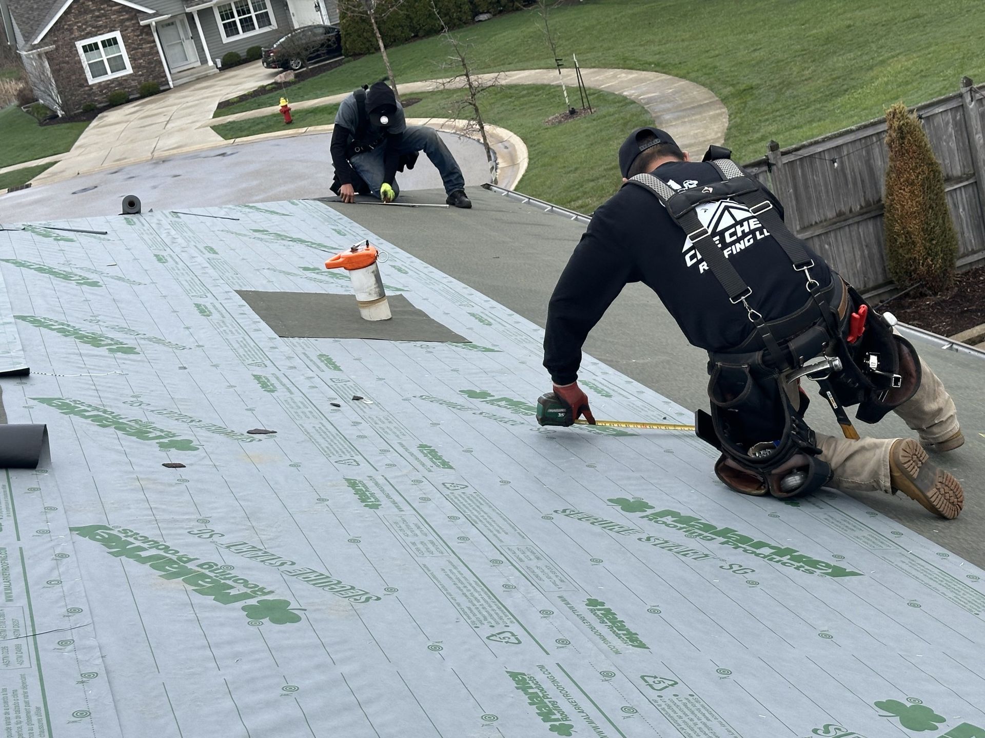 Two workers in safety gear install roofing underlayment on a slanted residential roof.