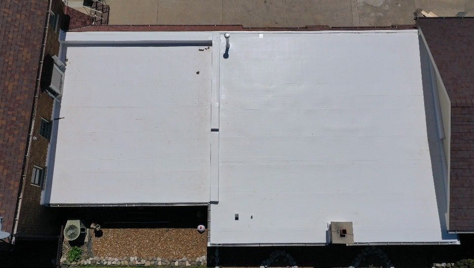 An aerial view shows a white flat roof section nestled between two traditional sloped, brown-shingled roof areas.